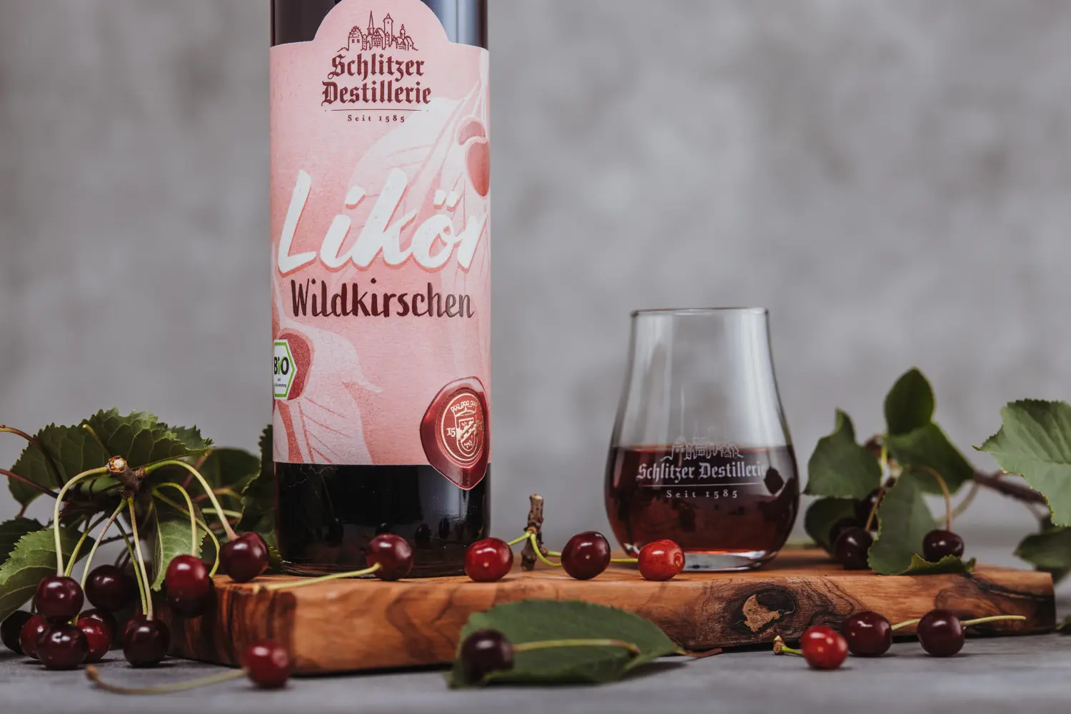 Wildkirschen Likör in eleganter Flasche – Fruchtiger Genuss aus der Schlitzer Destillerie Flasche Wildkirschlikör der Schlitzer Destillerie mit rotem Etikett, daneben frische Wildkirschen auf hellem Hintergrund – hochwertiger Fruchtlikör aus Deutschland.