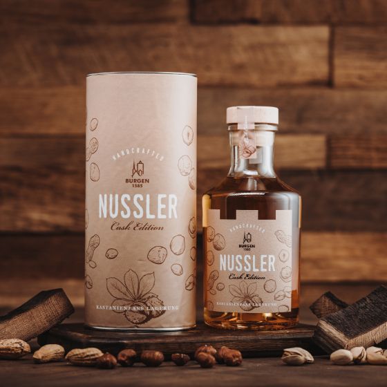 Schlitzer Nussler – Hochprozentiger Nusslikör in edler Flasche Flasche Schlitzer Nussler Haselnuss-Spirituose mit braunem Etikett und goldener Schrift auf hellem Hintergrund, daneben Haselnüsse und Holzlöffel zur Dekoration.