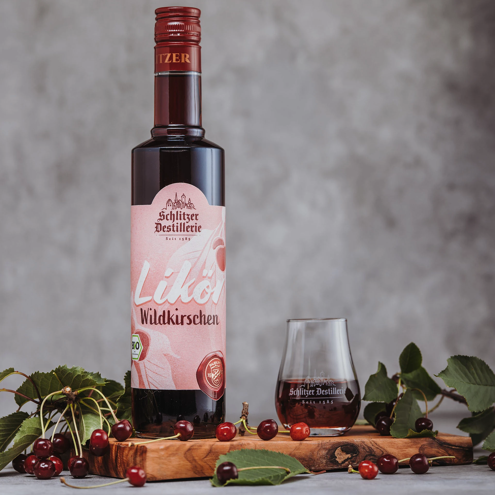 Schlitzer Wilkirschen Likör – Edler Fruchtlikör mit Wildkirschen in stilvoller Flasche Flasche Wilkirschen Likör der Schlitzer Destillerie mit rotem Etikett, umgeben von frischen Kirschen auf Holzuntergrund.