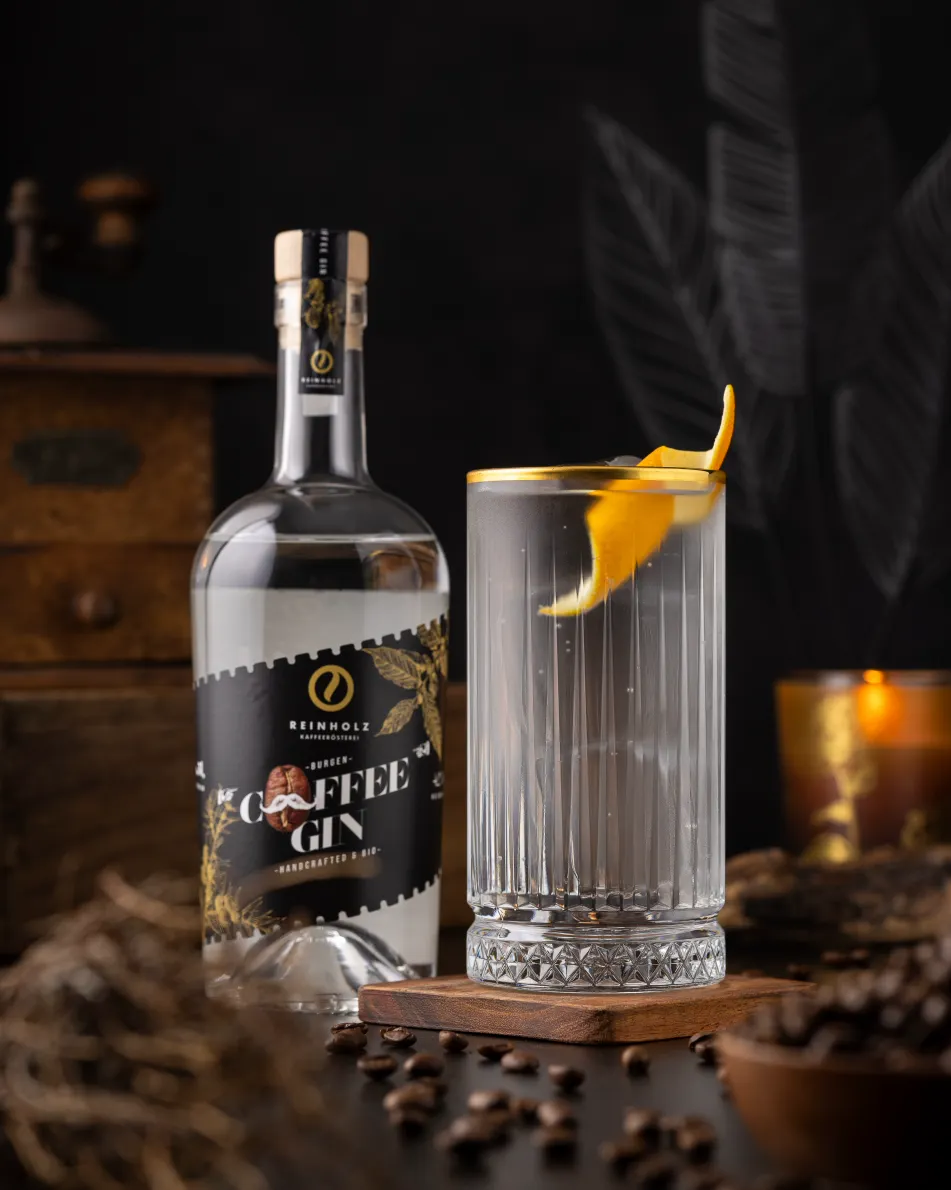 Kaffee Gin von der Schlitzer Destillerie – Aromatischer Kaffeegenuss trifft edlen Gin Braune 0,5-Liter-Flasche Schlitzer Kaffee Gin mit schwarzem Etikett, daneben Kaffeebohnen und ein Cocktail im Glas mit Eis, dekoriert mit Orangenzeste und Sternanis, vor hellem Hintergrund.