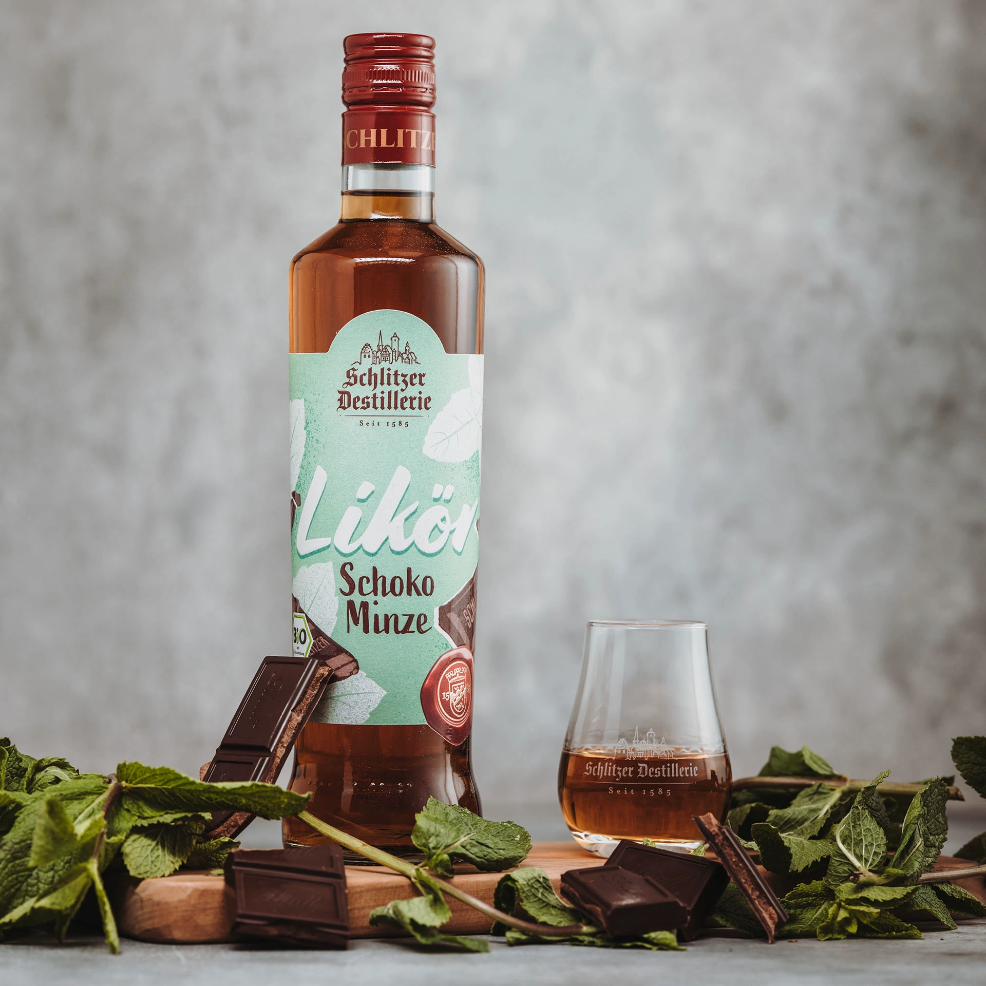Schlitzer Schoko-Minz Likör – Cremiger Minzlikör mit Schokoladennote in edler Flasche Flasche des Schoko-Minz Likörs der Schlitzer Destillerie mit braunem Etikett und Glaselementen, daneben ein Schnapsglas mit Likör und frische Minzblätter auf hellem Hintergrund.