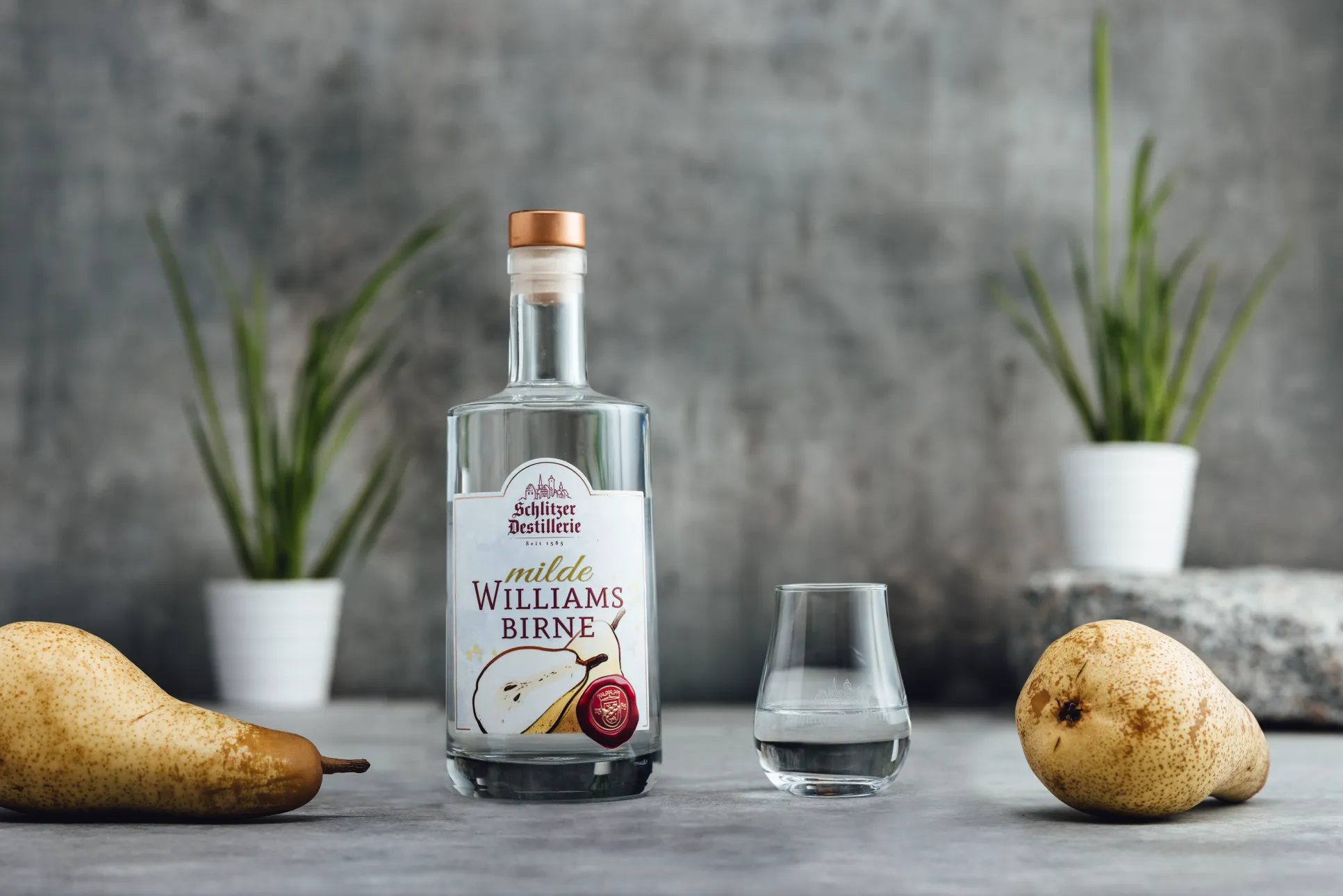 Milde Williams Birne – Edler Obstbrand aus Williams-Christ-Birnen | Schlitzer Destillerie Flasche Schlitzer Milde Williams-Birne Likör mit goldgelber Flüssigkeit, daneben reife Birnen und stilvolles Etikett mit Logo der Schlitzer Destillerie – Fruchtlikör aus Deutschland.