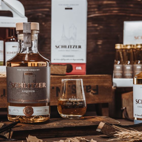 Deutscher Whisky in edlen Flaschen – Premium-Auswahl der Schlitzer Destillerie Drei Flaschen deutscher Whisky der Schlitzer Destillerie stehen auf einem rustikalen Holzfass; dunkel gehaltenes Ambiente mit stimmungsvollem Licht betont das edle Design der Flaschen und die goldbraune Farbe des Whiskys.