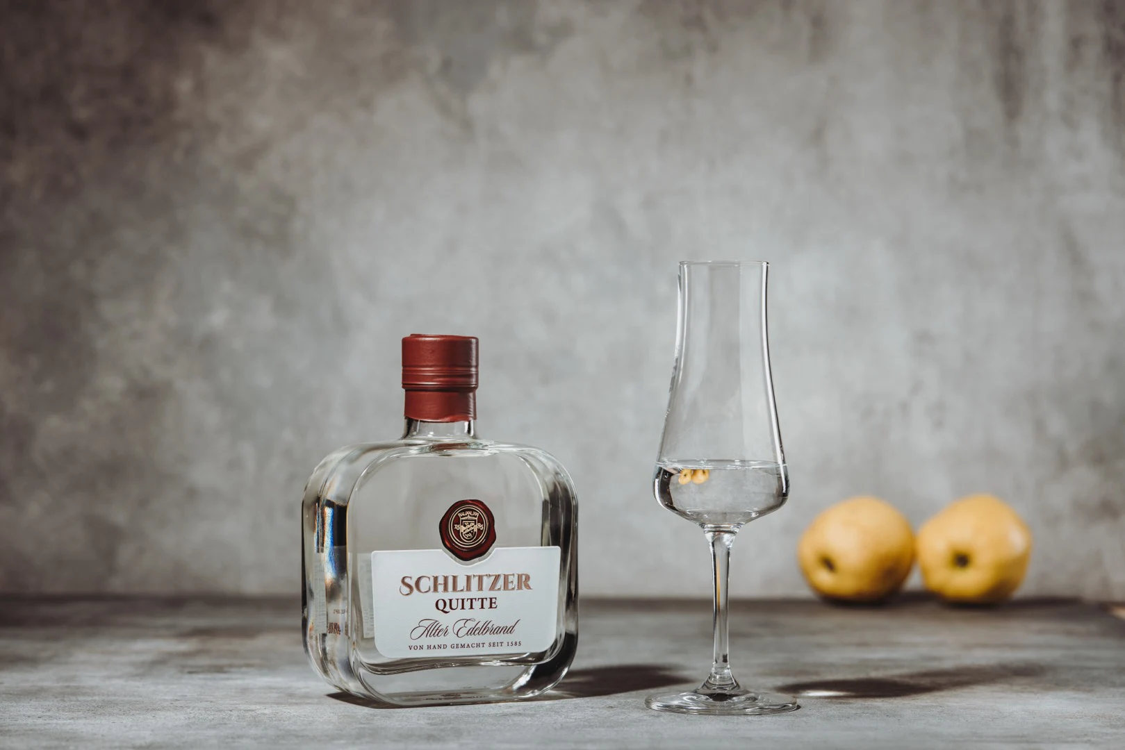Quitten Edelbrand – Hochwertiger Obstbrand aus sonnengereiften Quitten Quitten-Edelbrand in eleganter Flasche mit goldenem Etikett, auf Holzuntergrund inszeniert, umgeben von frischen Quitten – edler Obstbrand aus der Schlitzer Destillerie